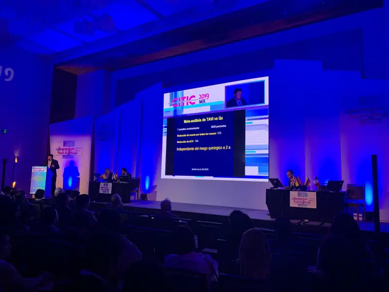 Congreso con ponente en el escenario, presentación en pantalla y público en la sala