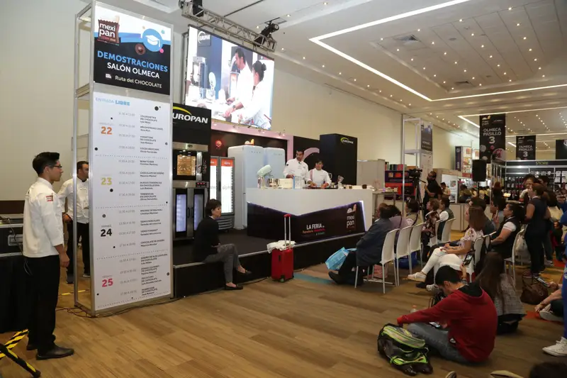 Escenario de demostraciones en una exposición, con pantalla, programa del día y público sentado